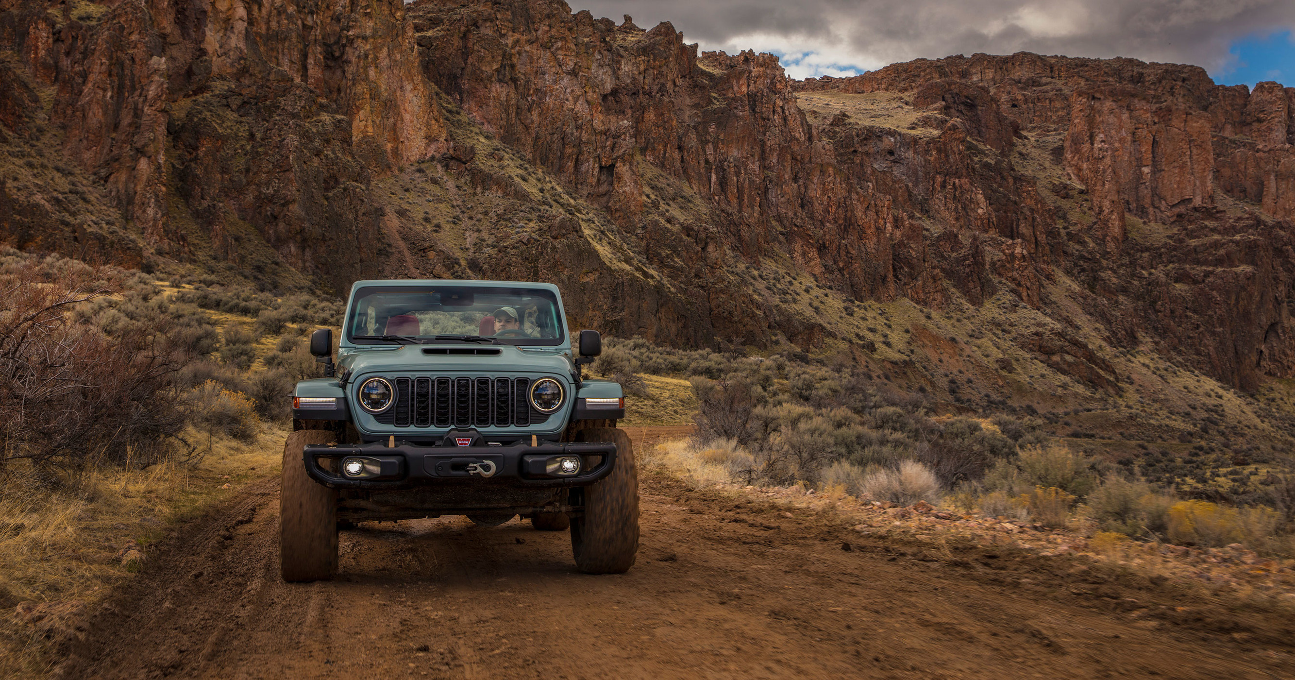 Front view of a grey 2024 Jeep Wrangler 4xe being driven down a desert road with the top down.