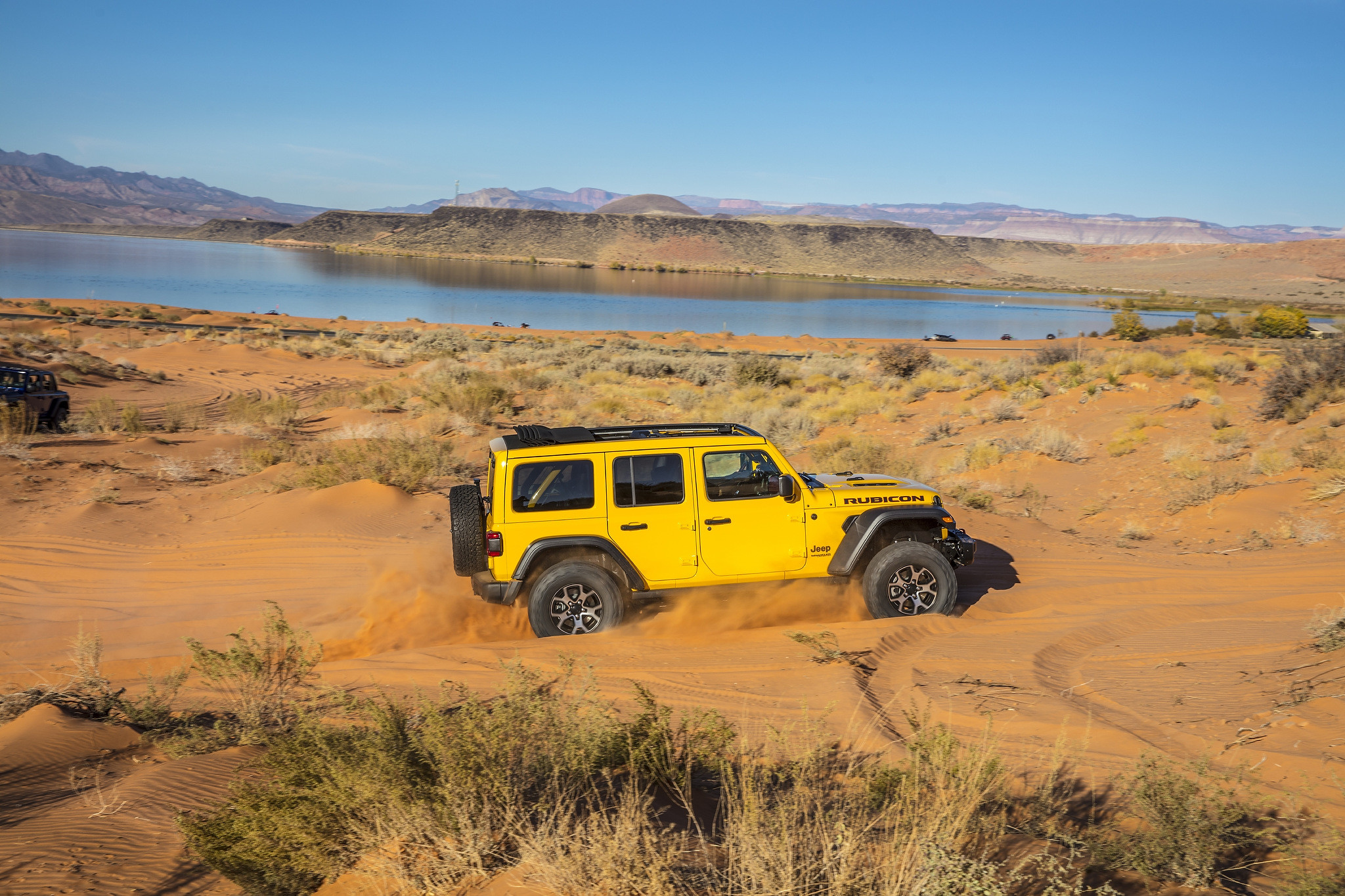 A 2021 Jeep Wrangler Rubicon with the roof open driving in a sand pit near a lake and followed by top-less 2021 Jeep Wrangler Rubicon
