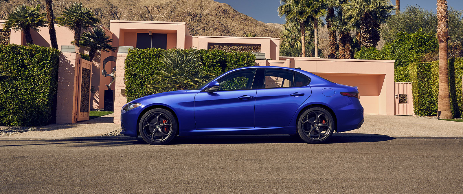 A side view of a blue 2021 Alfa Romeo Giulia parked in front of a pink bungalow surrounded by palm trees