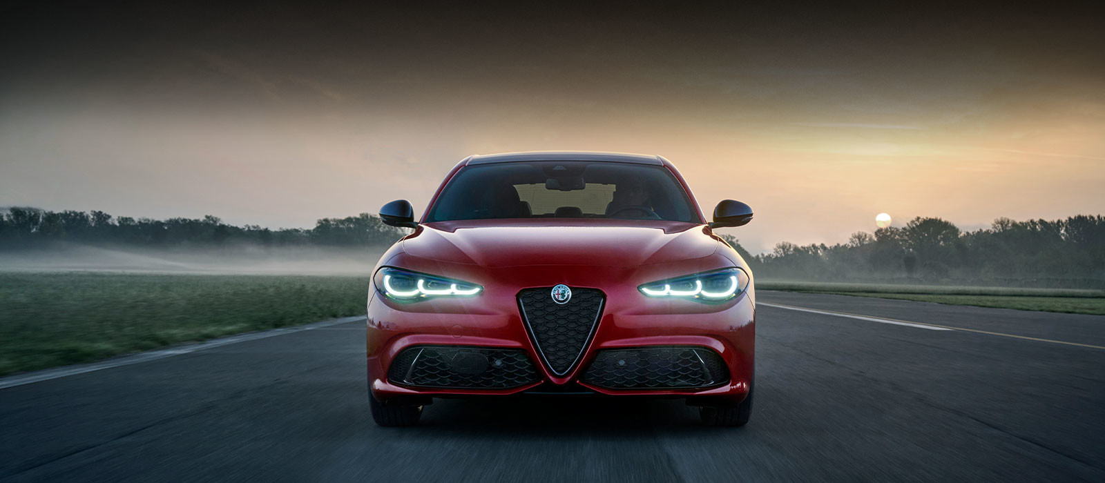 Vue de face d'une Alfa Romeo Giulia 2025 couleur Alfa Rosso, sur une route au lever du soleil, avec un paysage brumeux et des arbres au loin.