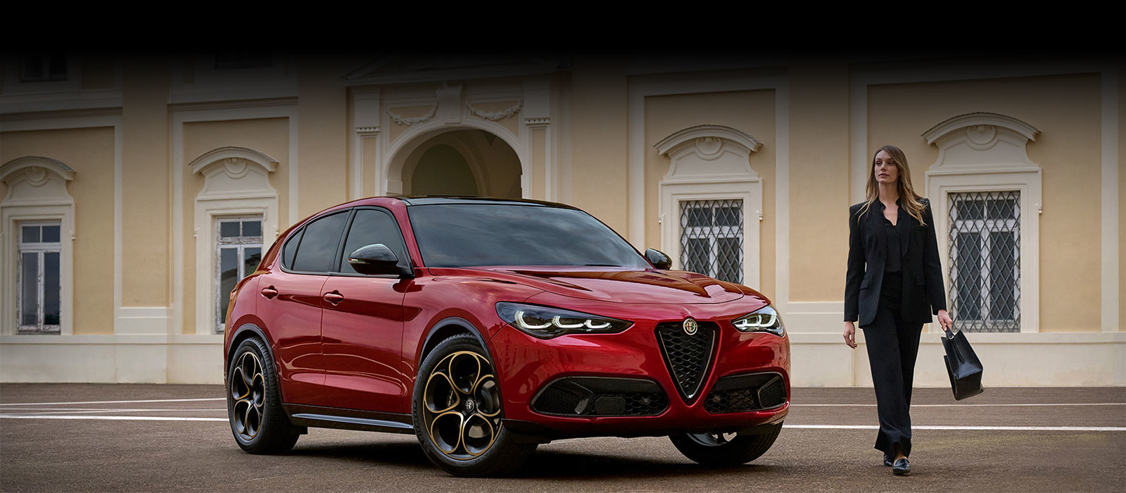 Un VUS Alfa Romeo rouge est stationné devant un bâtiment classique avec des fenêtres en cintre. Une personne vêtue de noir et portant un sac à main marche à côté de la voiture.
