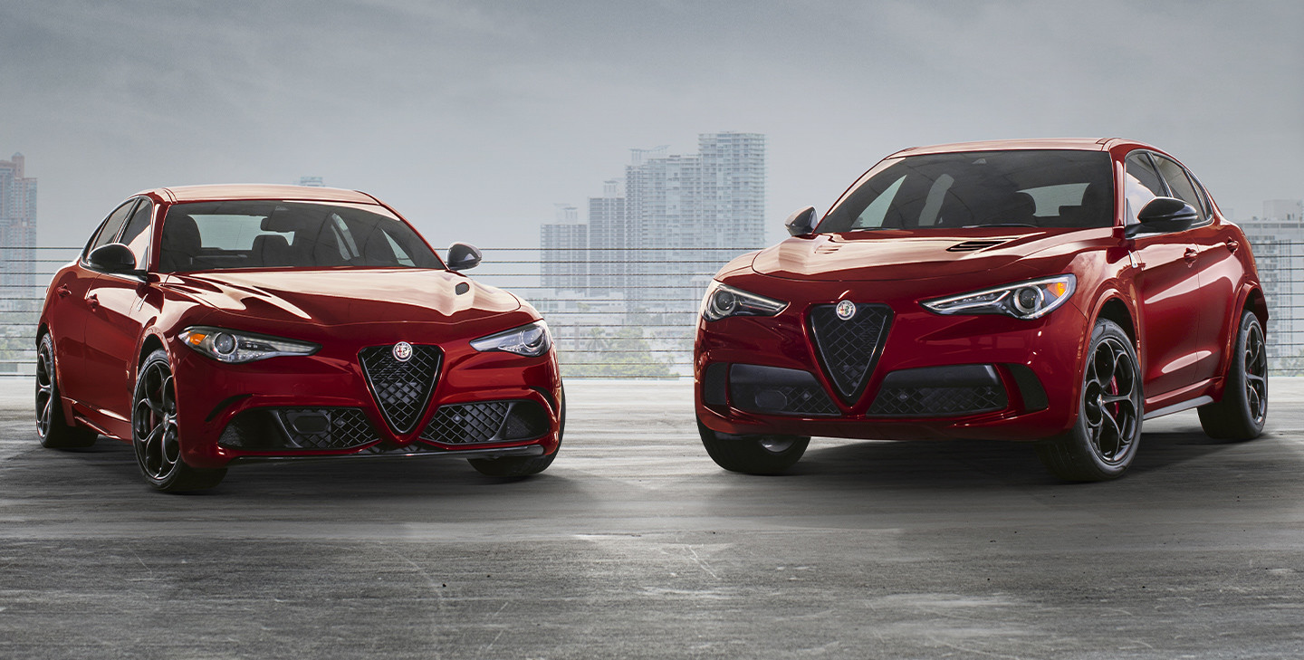 An Alfa Romeo Giulia and an Alfa Romeo Stelvio, both in Rosso GTA, both parked in a parking lot with a city skyline in the background.