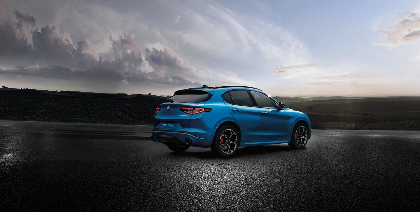 La vue arrière d'une Alfa Romeo Stelvio 2023 bleue montrée garée sur un trottoir avec des arbres et un ciel rempli de nuages au loin.