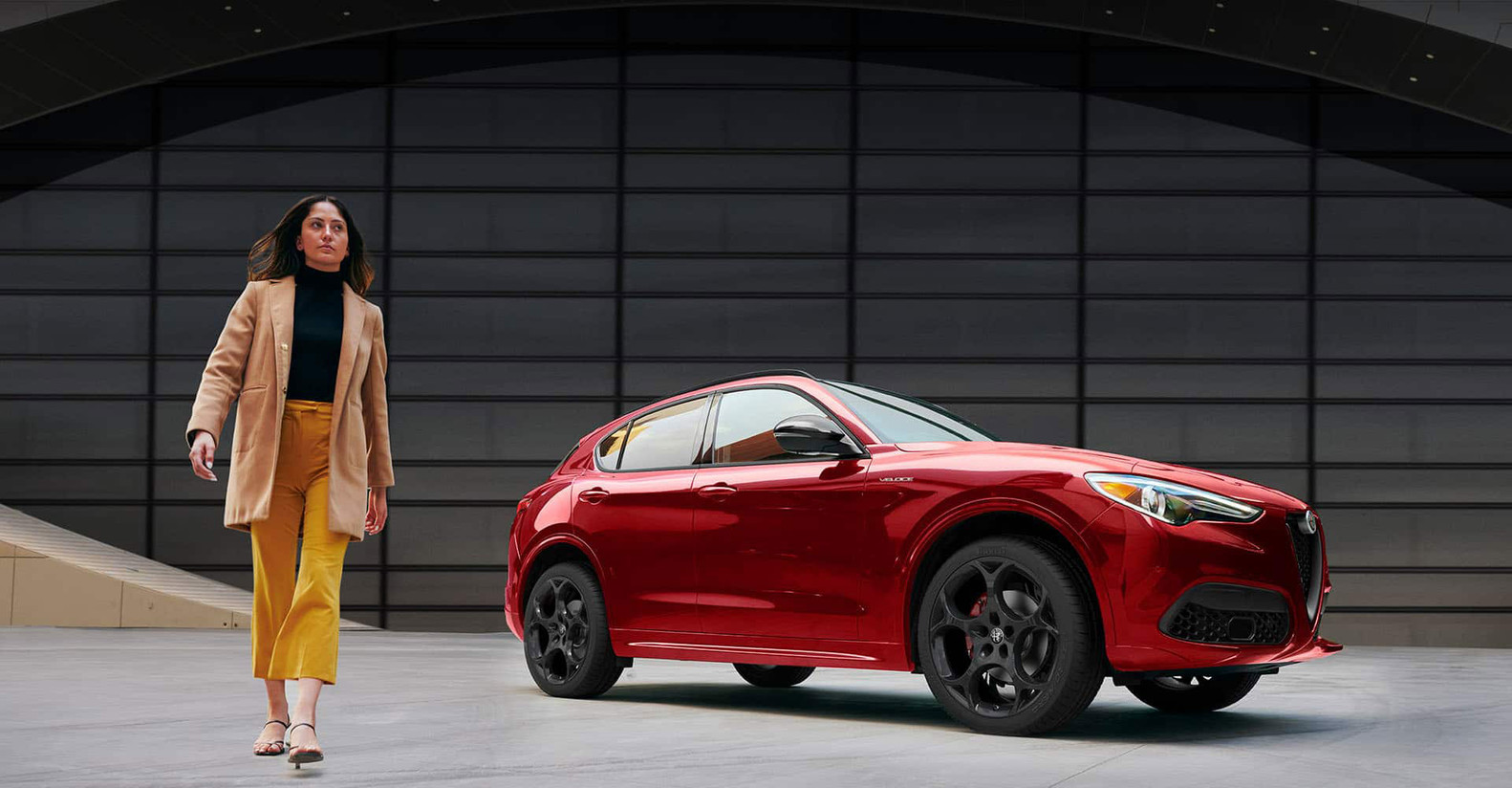 A red Alfa Romeo Stelvio, parked in front of a building, with a person walking away from the car.
