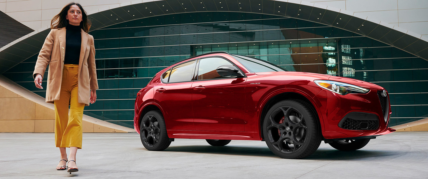 A woman struts next to her new red Alfa Romeo 2022 Stelvio parked in front of a concrete building