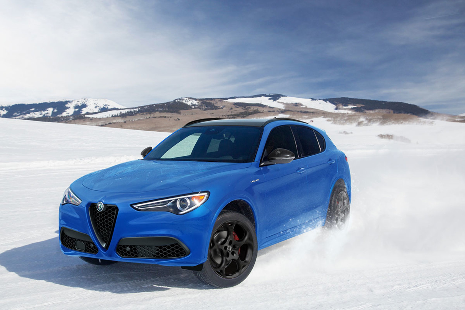 Vue latérale avant d’un Alfa Romeo Stelvio 2023 rouge roulant sur une route enneigée avec des montagnes en arrière-plan.