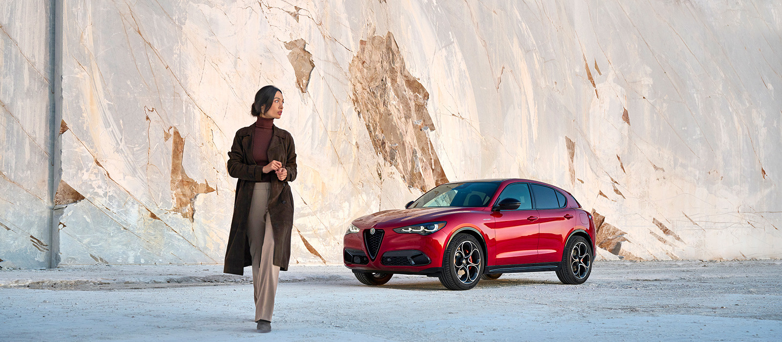 Vue d'une Alfa Romeo Stelvio 2024 rouge garée sur un fond en forme de rocher et d'une femme qui s'éloigne du véhicule.