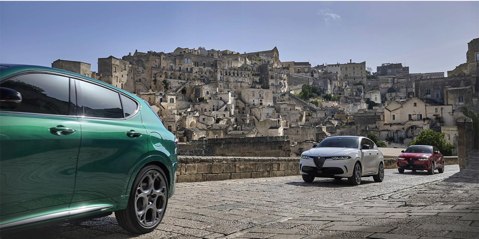 Trois voitures Alfa Romeo Tonale Tributo Italiano, de couleur respectivement verte, blanche et rouge, roulant sur une route en béton, avec des bâtiments de style italien en arrière-plan.