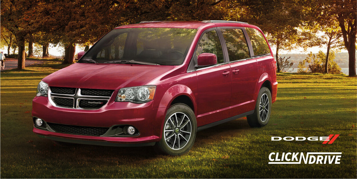 Dodge Click ’n Drive. Red Dodge Grand Caravan parked on grass with trees and water in the background.