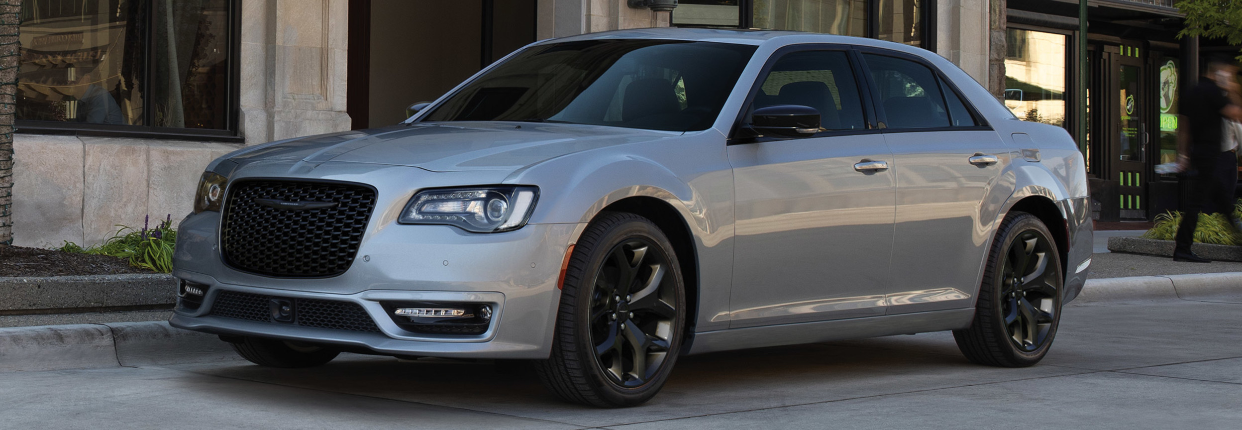 Vue latérale d’une Chrysler 300 2021 argent éclatant, garée devant l’entrée d’un immeuble en copropriété.