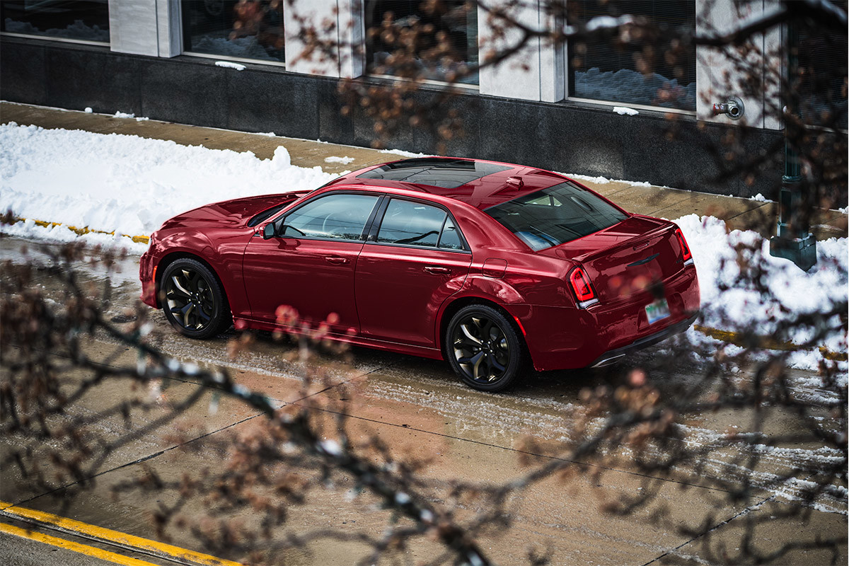 An elevated side view of a Velvet Red Pearl 2021 Chrysler 300 parked on a road next to a snow-covered sidewalk.