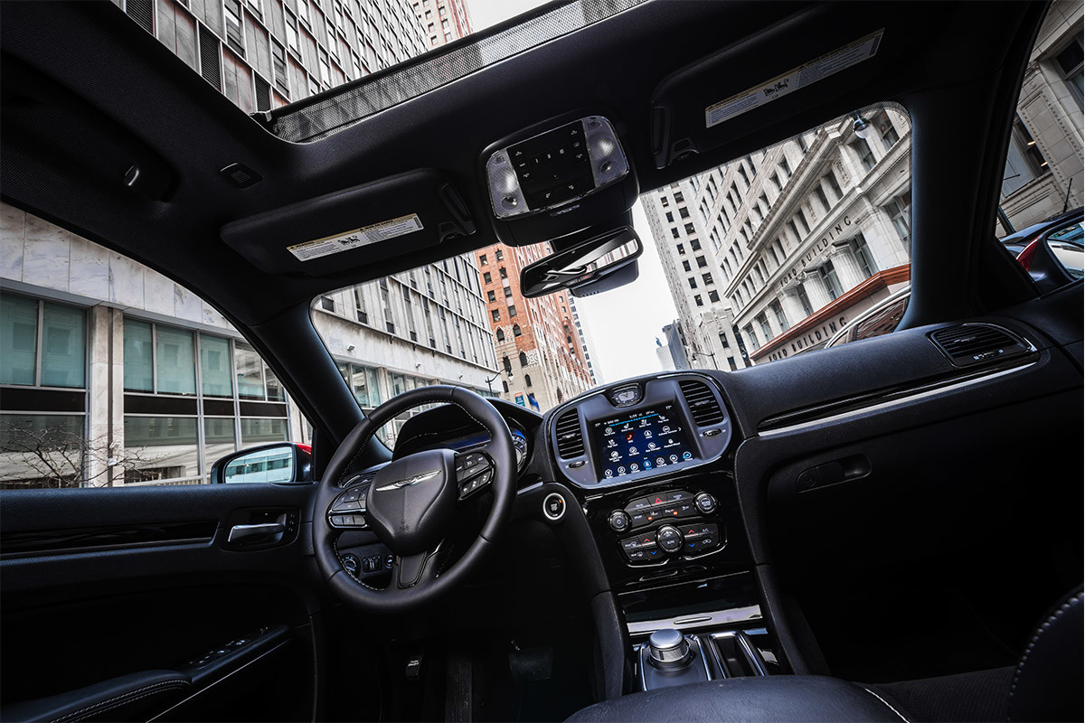 Vue intérieure de l’habitacle élégant de la Chrysler 300 2021. Le toit ouvrant panoramique à deux panneaux, livrable en option, est ouvert, révélant les gratte-ciel de la ville au-dessus de lui.