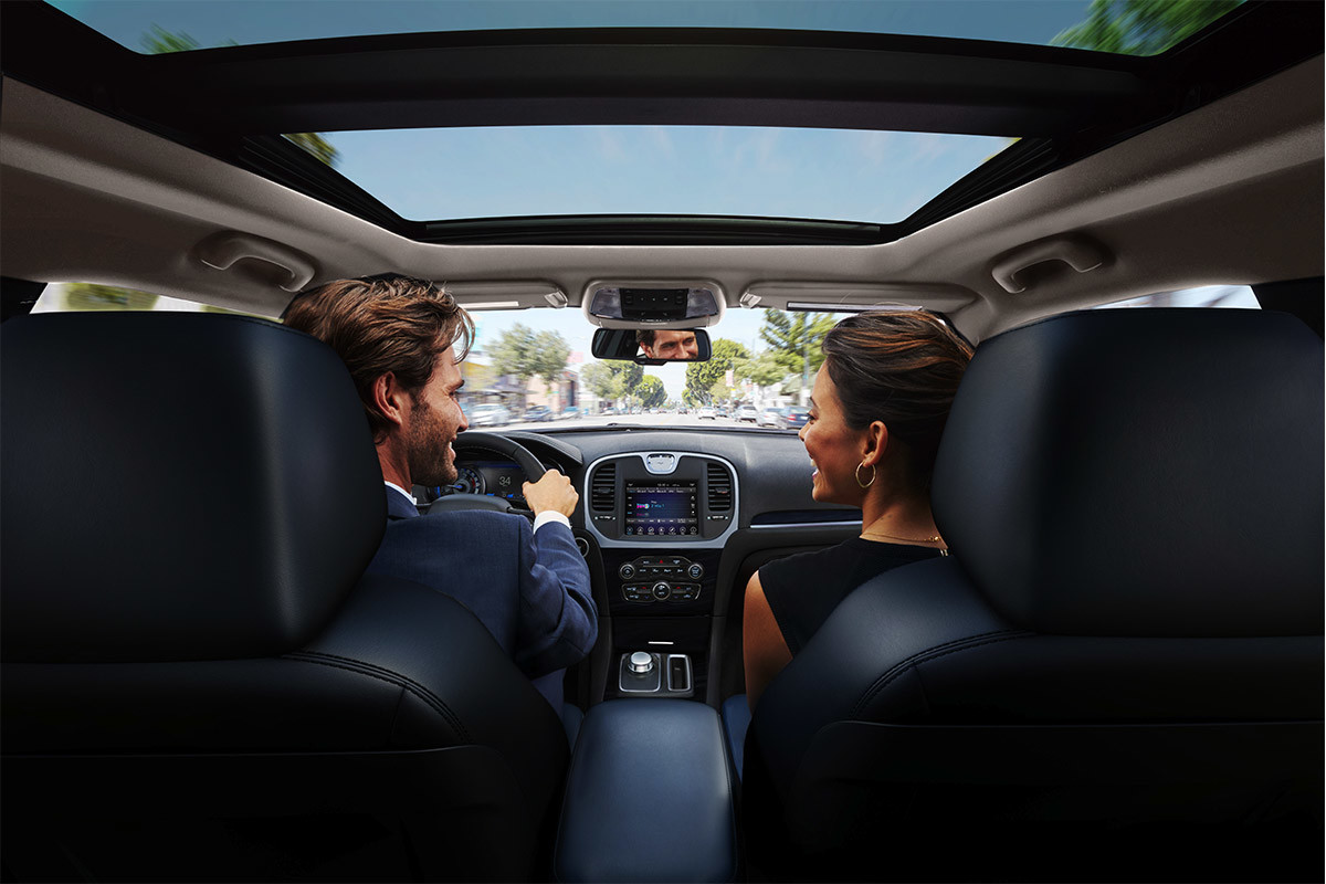 Un homme et une femme assis sur les sièges avant à dessus en cuir noir de la Chrysler 300 2021, en train de discuter avec le toit ouvrant panoramique à deux panneaux, livrable en option, ouvert au-dessus d’eux.