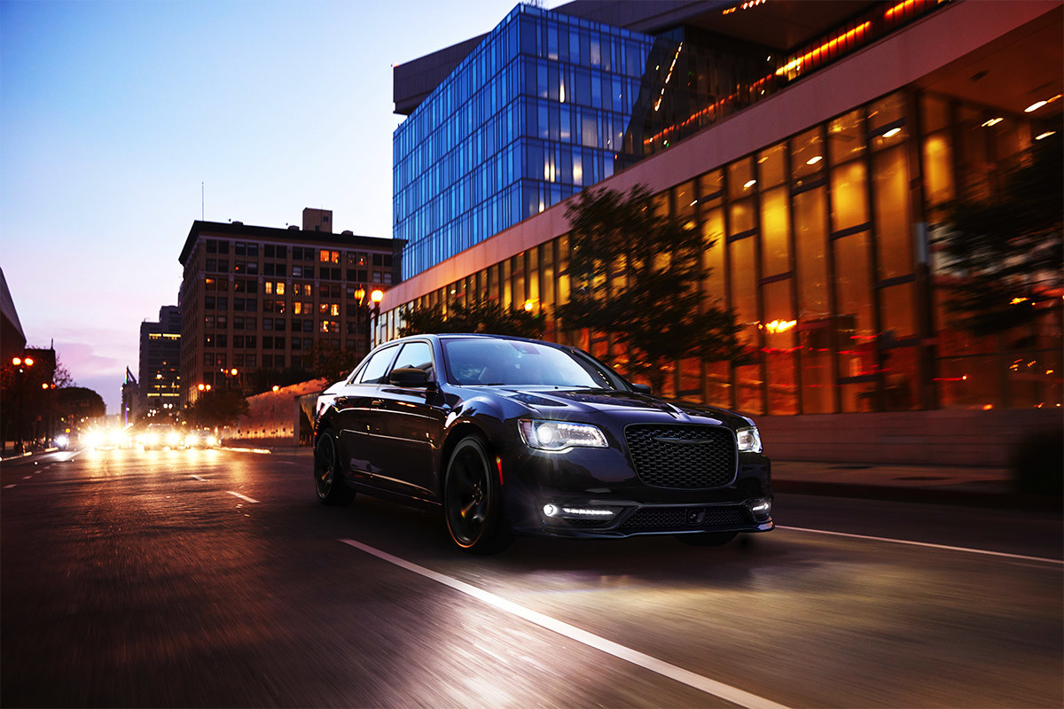 Vue de face d’une Chrysler 300 2021 noir brillant roulant de nuit sur une route urbaine. Les phares et les antibrouillards à DEL du véhicule brillent de tous leurs feux.