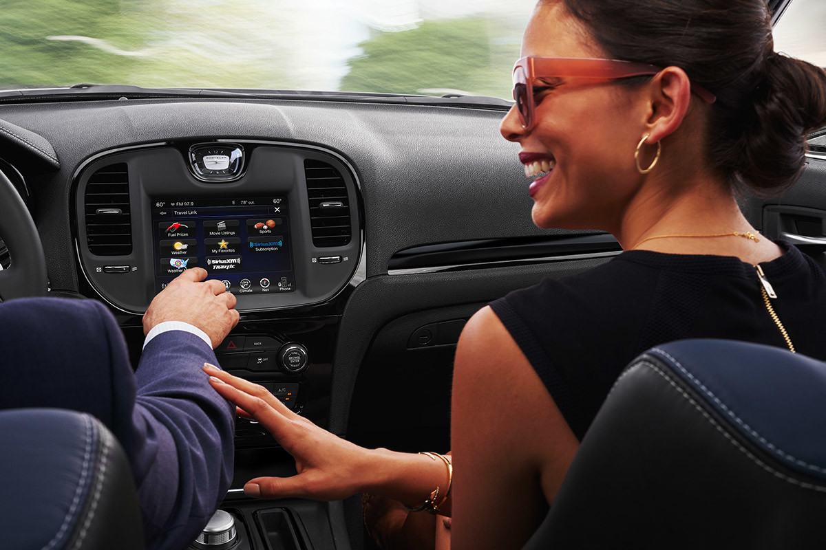Gros plan d’une femme souriante sur le siège passager avant d’une Chrysler 300 2021. Elle touche le bras du conducteur alors qu’il sélectionne une fonction du centre multimédia Uconnect à écran tactile de 8,4 po.