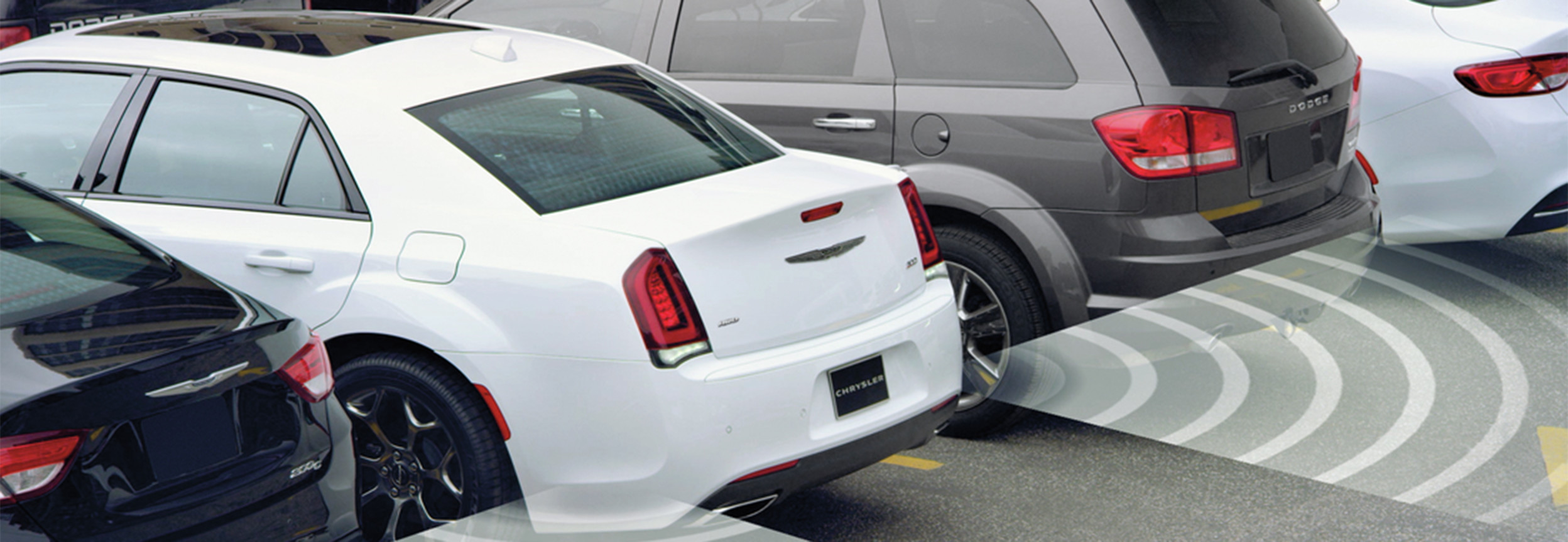 An elevated view of a Bright White Chrysler 300 parked in a parking space. Illustrated arcs emanate from the vehicle’s rear corners to indicate the range of the ParkView Rear Back Up Camera and Park-Sense System.