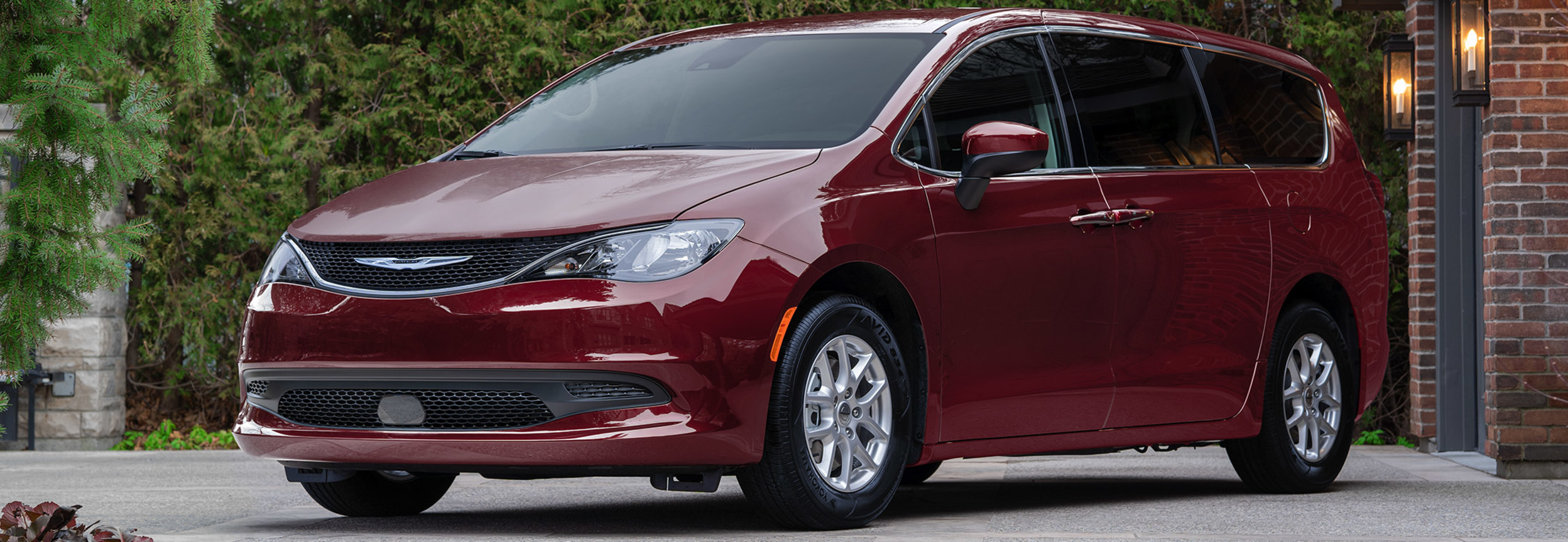 A three-quarters view of a Velvet Red Pearl 2021 Chrysler Grand Caravan parked on the driveway of a red-bricked house.