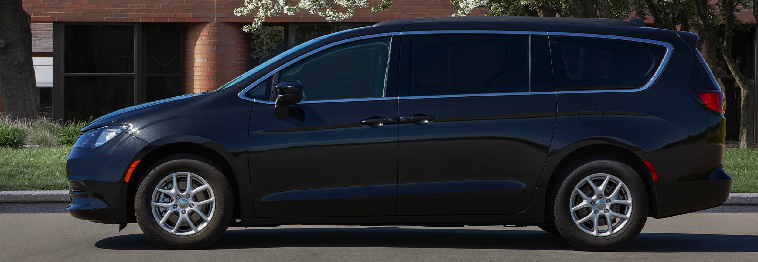 A three-quarter view of the athletic exterior of a Granite Crystal Metallic 2021 Chrysler Grand Caravan parked on a road in front of a red-bricked college.
