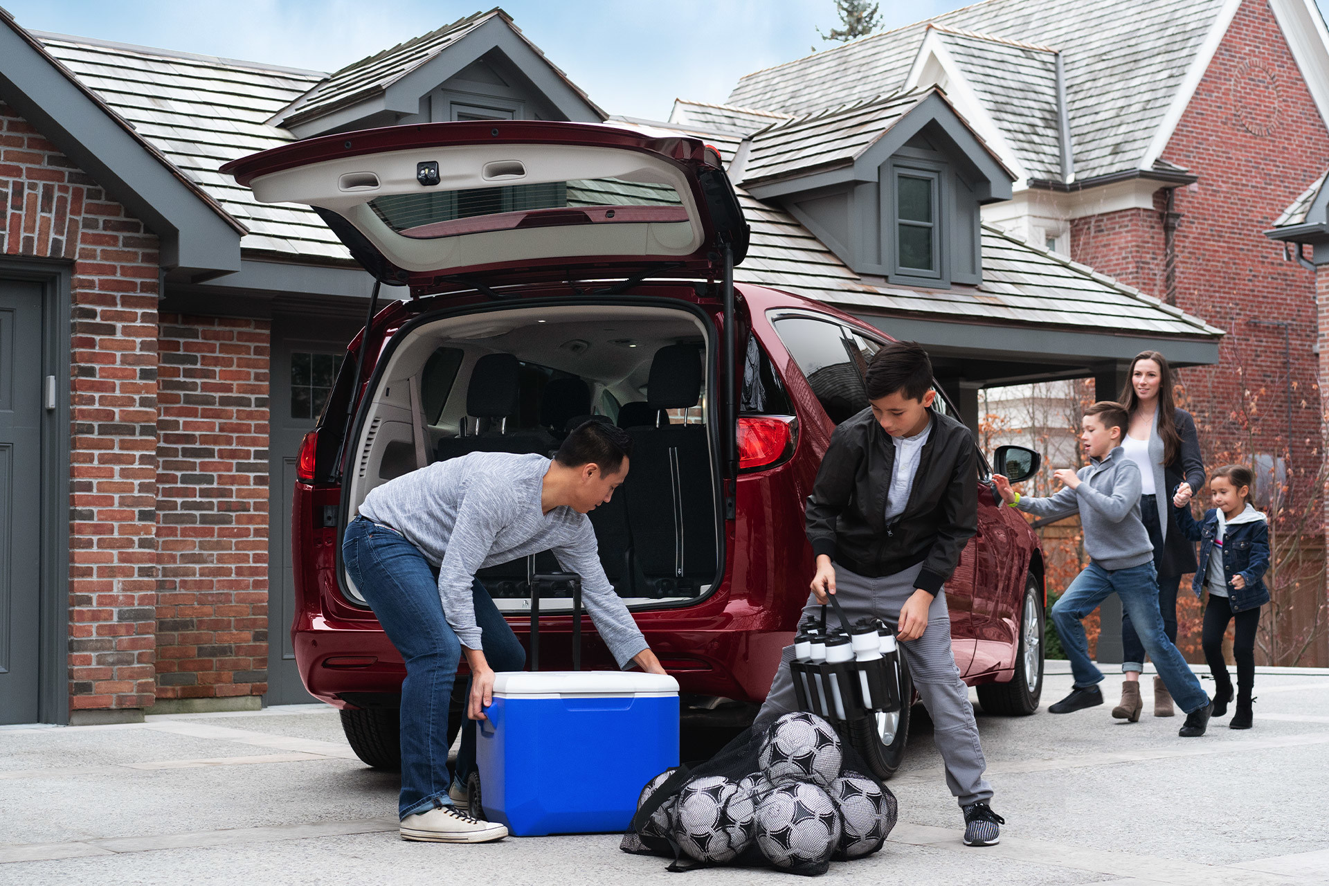 Vue arrière d’un homme et un garçon sortant une glacière et un sac de ballons de soccer d’une Chrysler Grand Caravan 2021 rouge nacrée de velours, garée devant une maison de briques rouges avec des enfants qui jouent à l’arrière