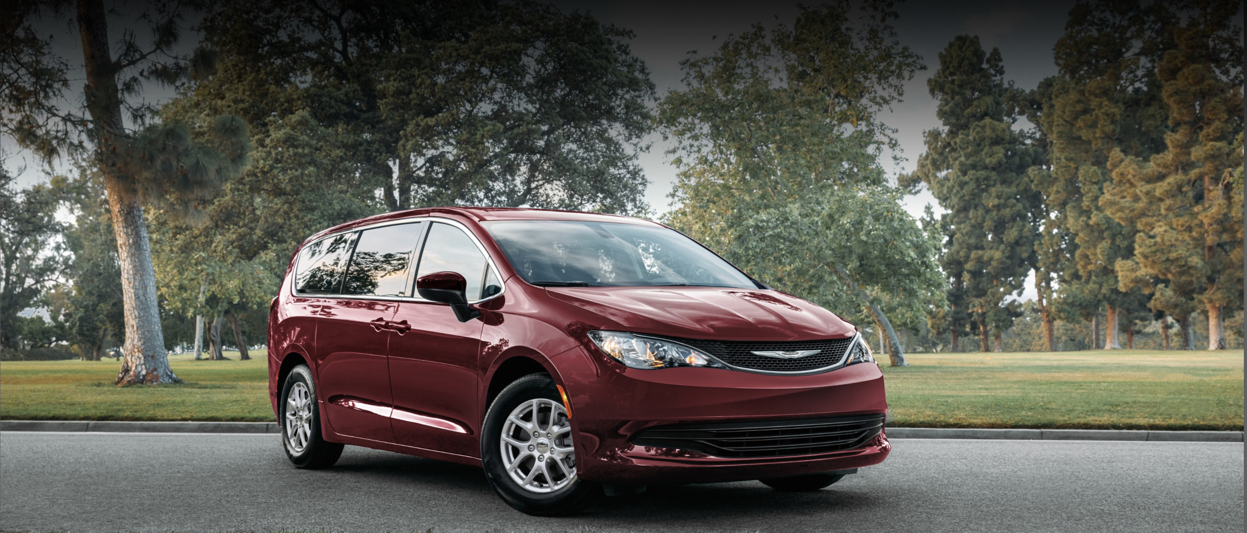The new 2021 Chrysler Grand Caravan in red, parked on a paved road surrounded by grass and trees.