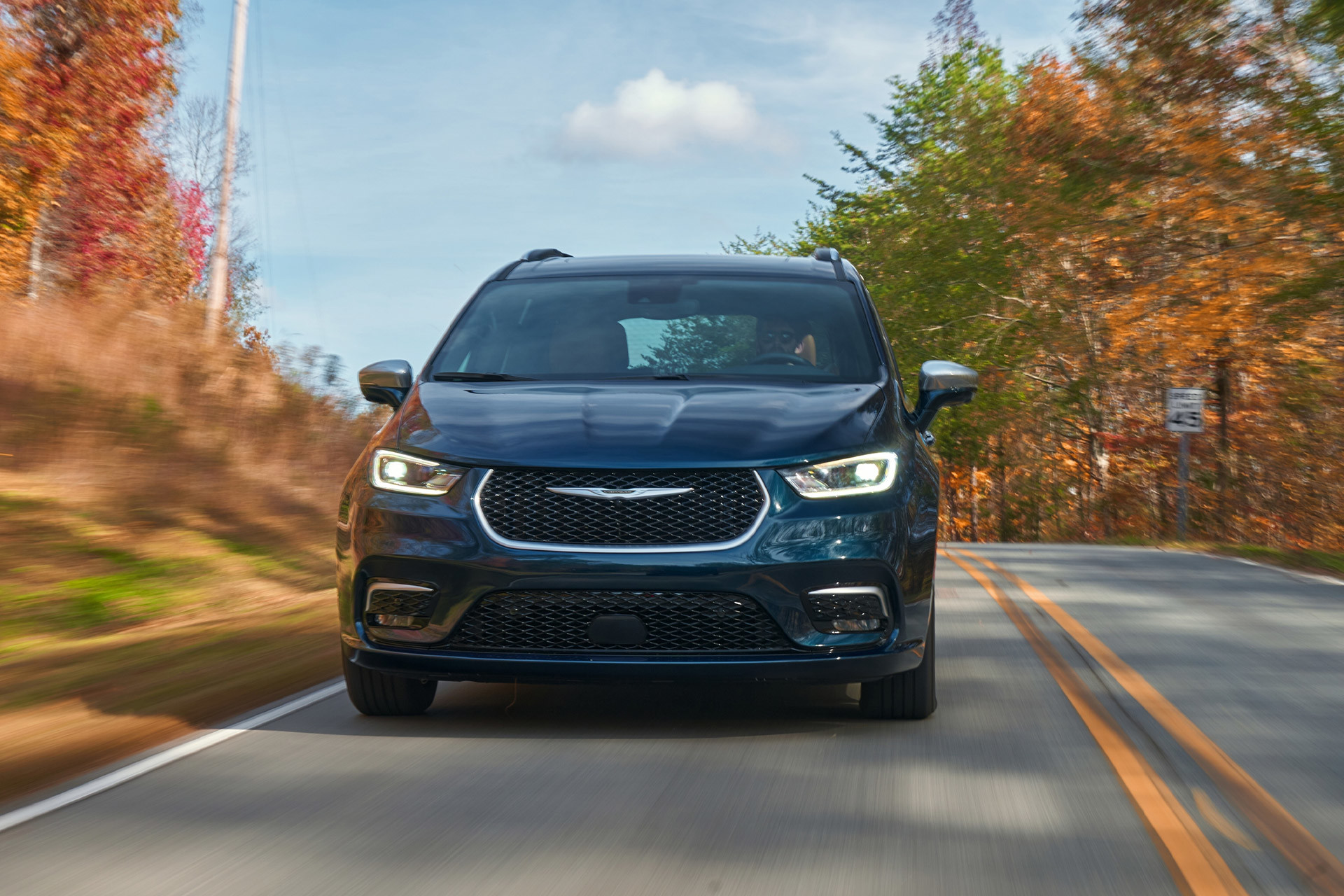 Front view of a teal 2023 Chrysler Electric vehicle driving down a paved road with forest scenery in the background.