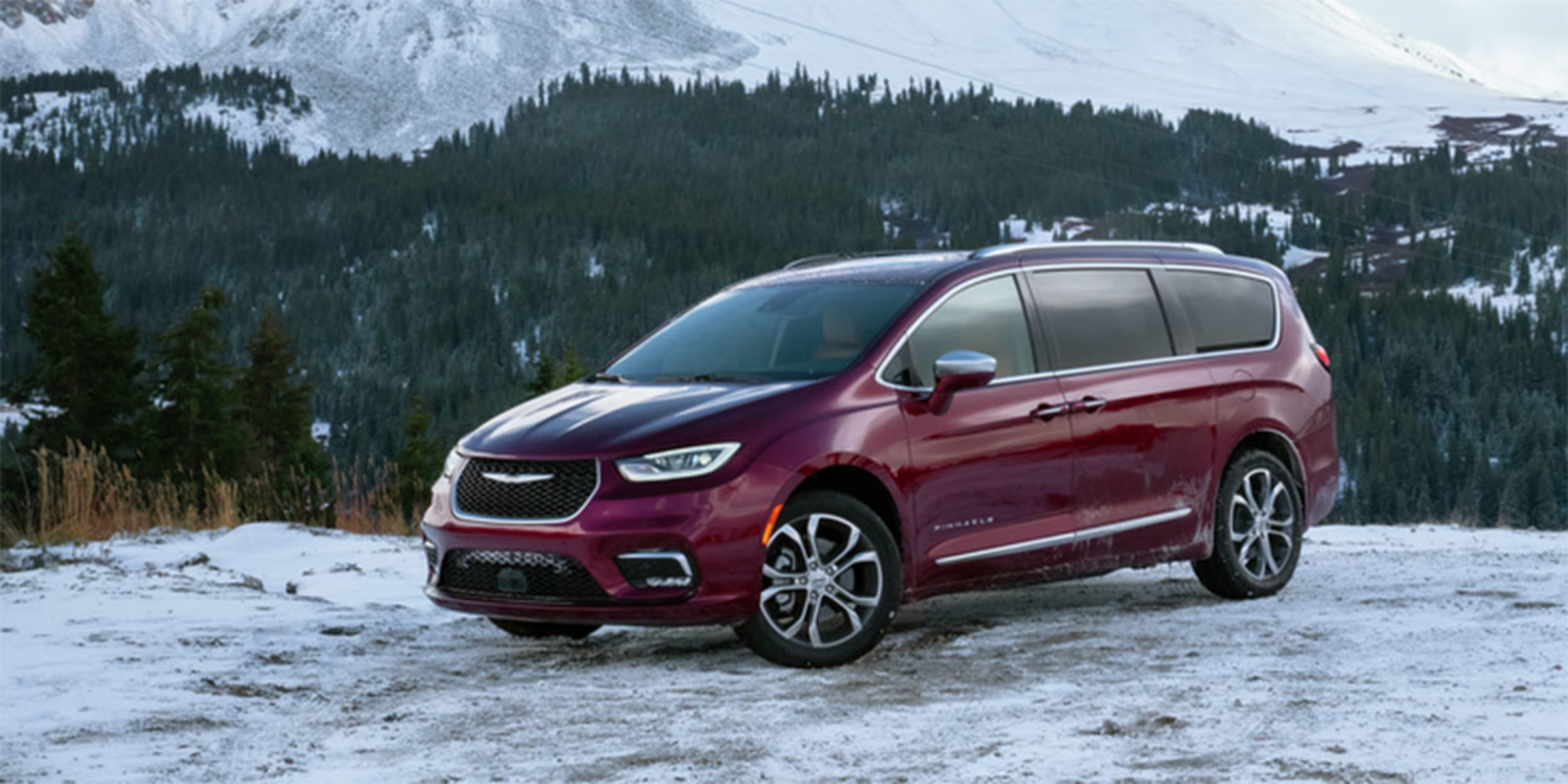 View of a red 2023 Chrysler Pacifia parked on snow covered ground with mountains and trees behind.