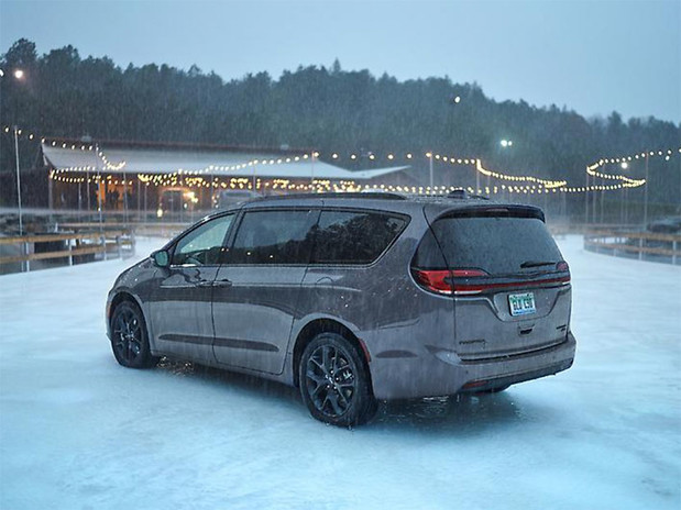Vue d'une Chrysler Pacifica 2023 gris foncé garée à l'extérieur d'une cabane en rondins orange avec des arbres couverts de neige en arrière-plan.