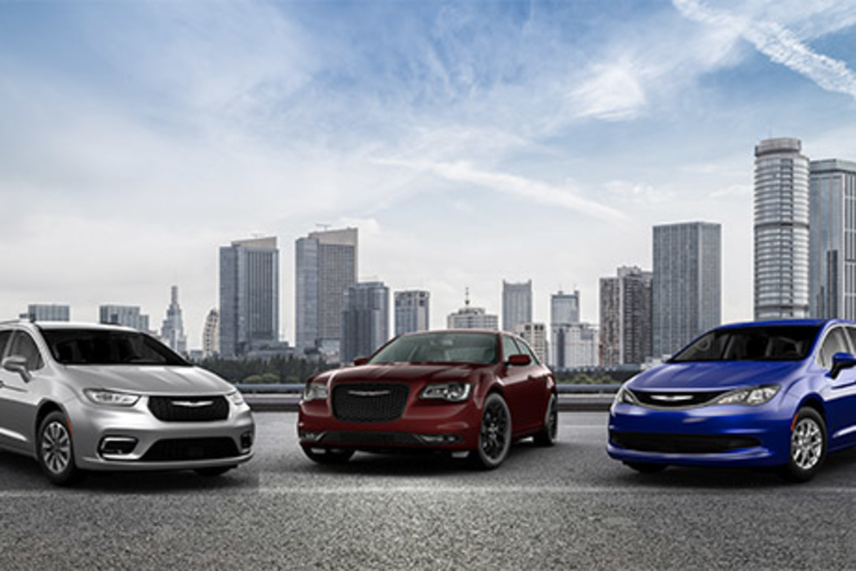 Une Chrysler Pacifica grise, une Chrysler 300 rouge et une Chrysler Grand Caravan bleue, toutes garées sur une route pavée avec une ligne d’horizon de la ville en arrière-plan.