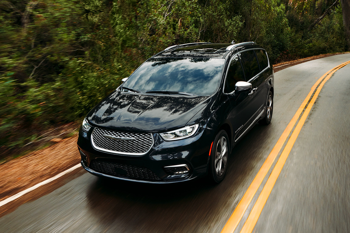 The new 2021 Chrysler Pacifica in black driving down a road with forest on both sides.