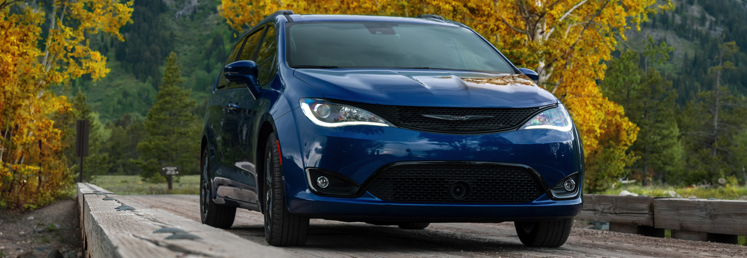 Chrysler Pacifica bleue roulant sur un petit pont entouré de montagnes et d’arbres aux couleurs d’automne.