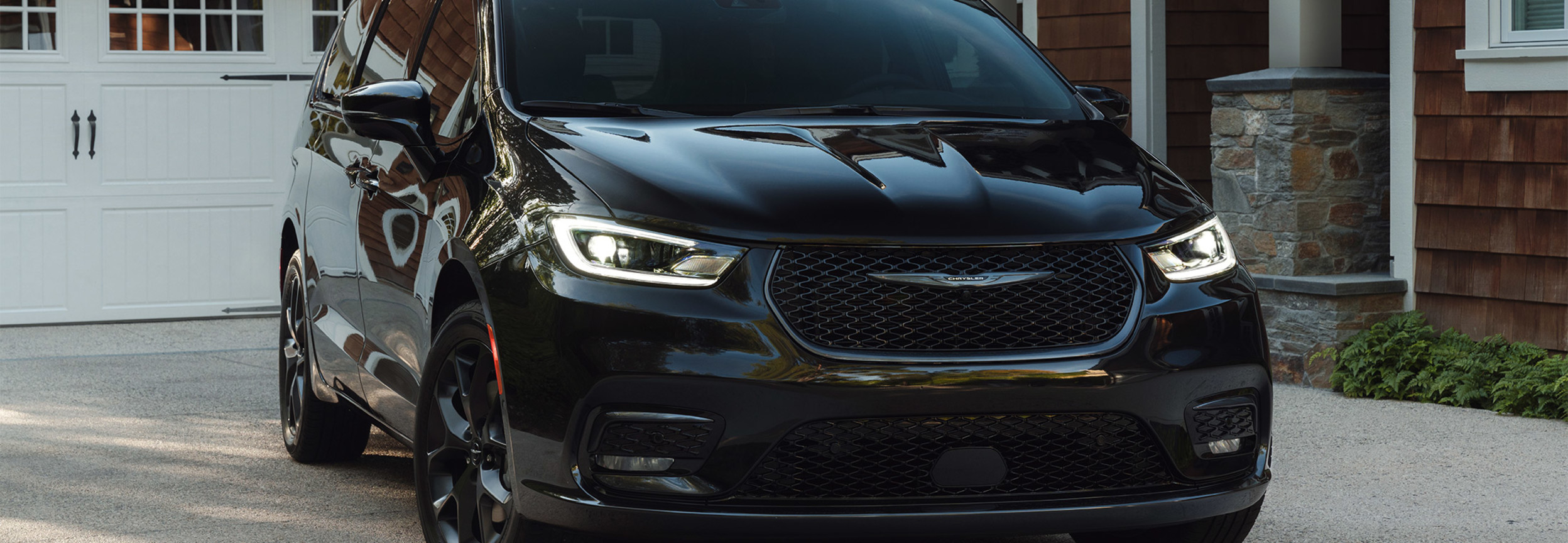 A front view of a Brilliant Black Crystal Pearl 2021 Chrysler Pacifica parked in the driveway of stylish wood-panelled home.