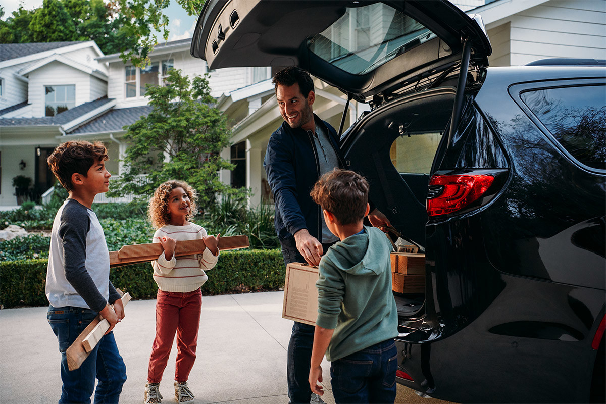 A view of three children helping a man unload items from the cargo area of a Brilliant Black Crystal Pearl 2021 Chrysler Pacifica.