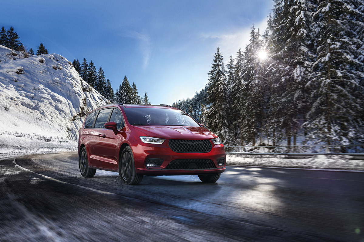 A front view of a Velvet Red Pearl 2021 Chrysler Pacifica being driven on a mountain road as the sun peeks through snow-covered trees.