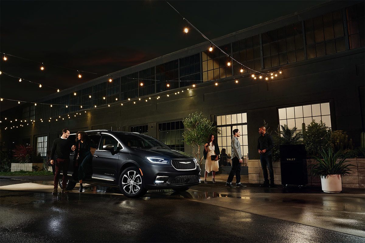 A view of a man and a woman leaning on the open sliding door of a Maximum Steel Metallic 2021 Chrysler Pacifica as people socialize on a patio beside the vehicle.