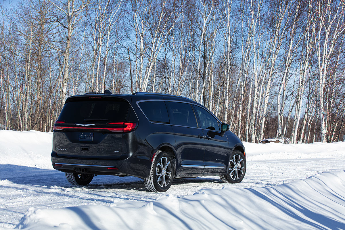 A rear view of a Brilliant Black Crystal Pearl 2021 Chrysler Pacifica parked on a snow-covered road with birch trees in the background.
