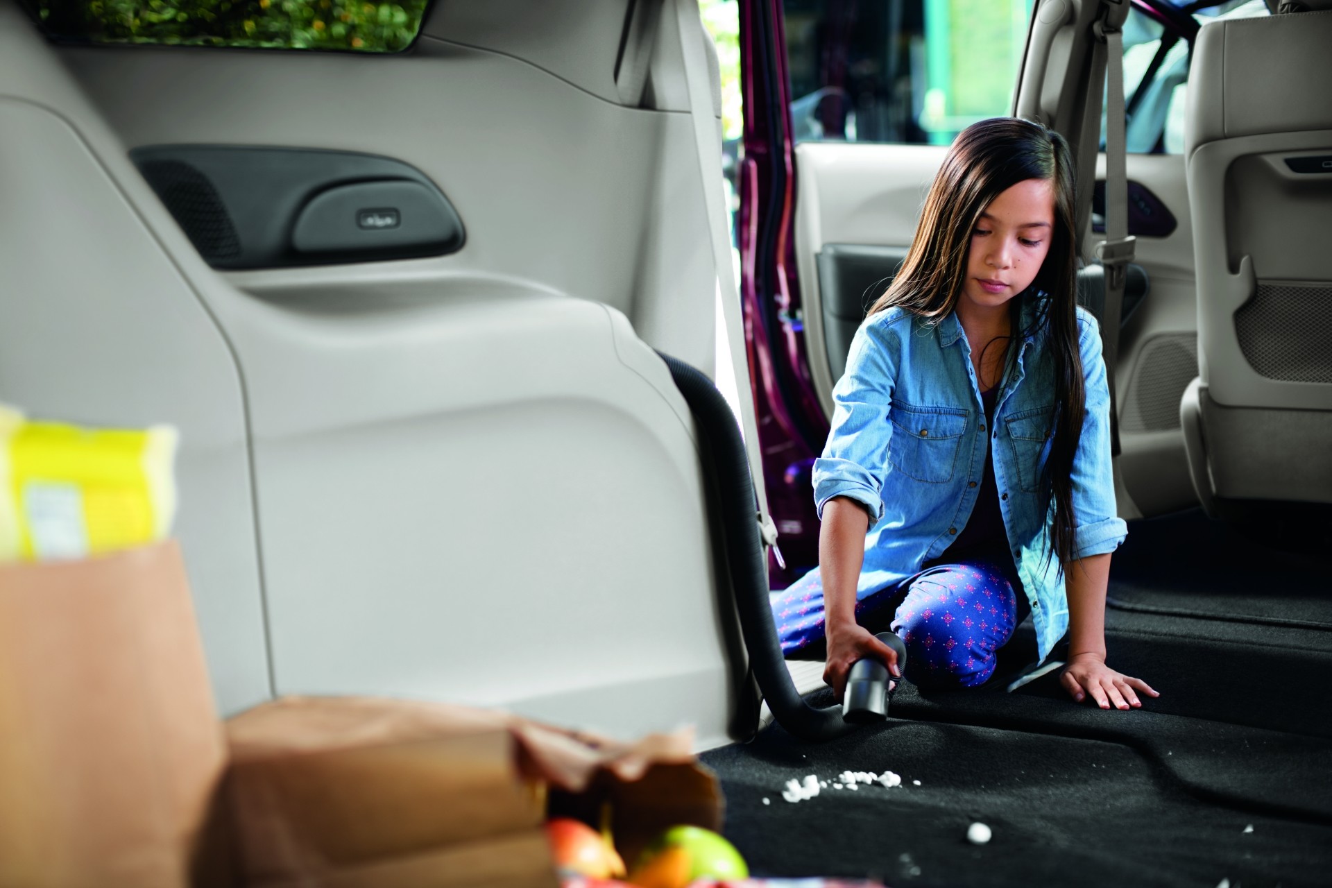 Vue d’une fille assise sur le plancher d’une Chrysler Pacifica en train de ramasser quelques miettes avec l’aspirateur intégré Stow n’ Vac livrable en option.