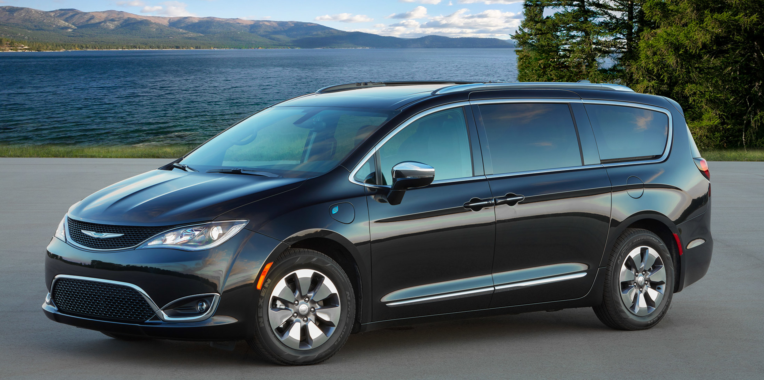 A black Chrysler Pacifica hybrid, parked on a paved road with a lake, trees, and mountains in the background.