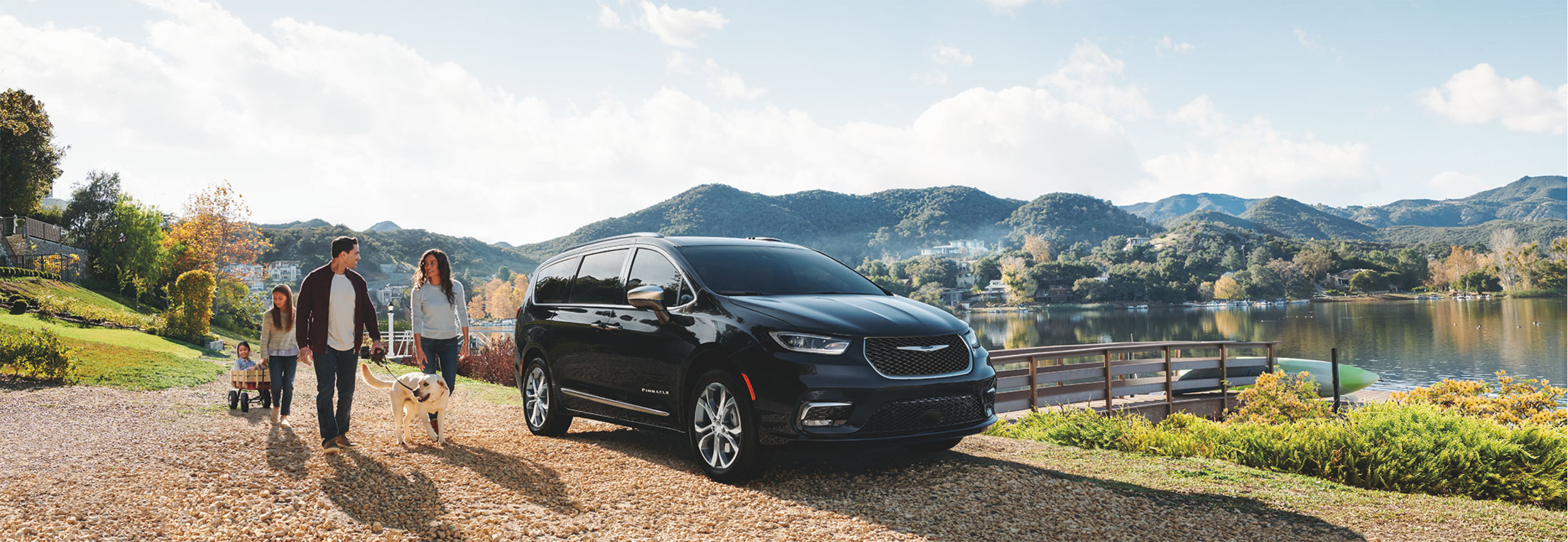 A black 2022 Chrysler Pacifica Hybrid, parked in front of a lake with a family of four walking their dog in the background.