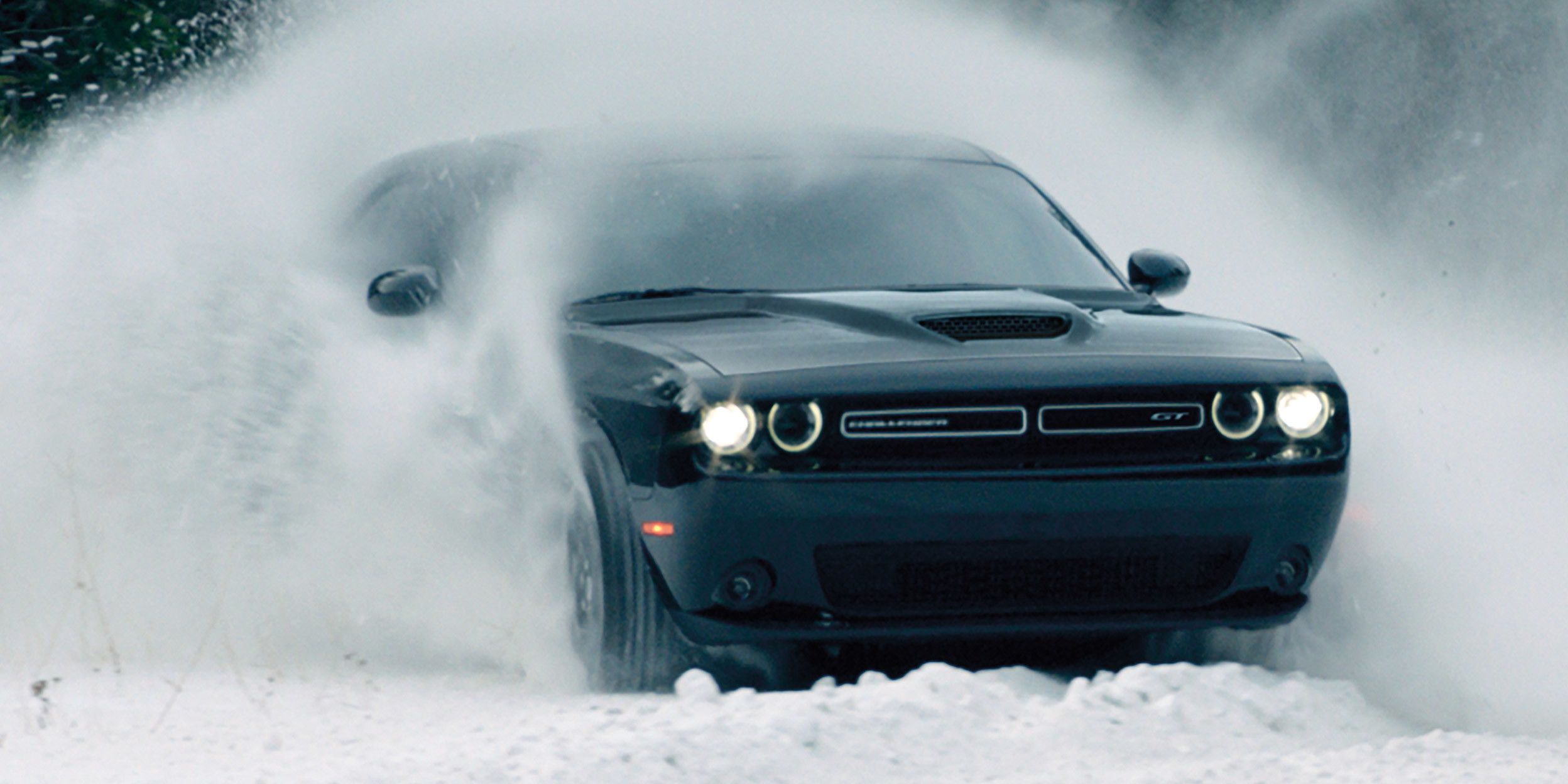 Challenger SXT AWD in Black driving through snowy field on overcast day with headlamps on and snow flying up around vehicle.