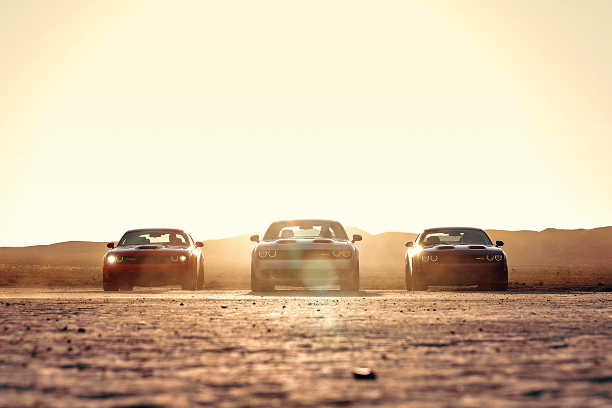 Three 2021 Dodge Challenger muscle cars parked on a desert plain as the sun rises over the hills behind them.
