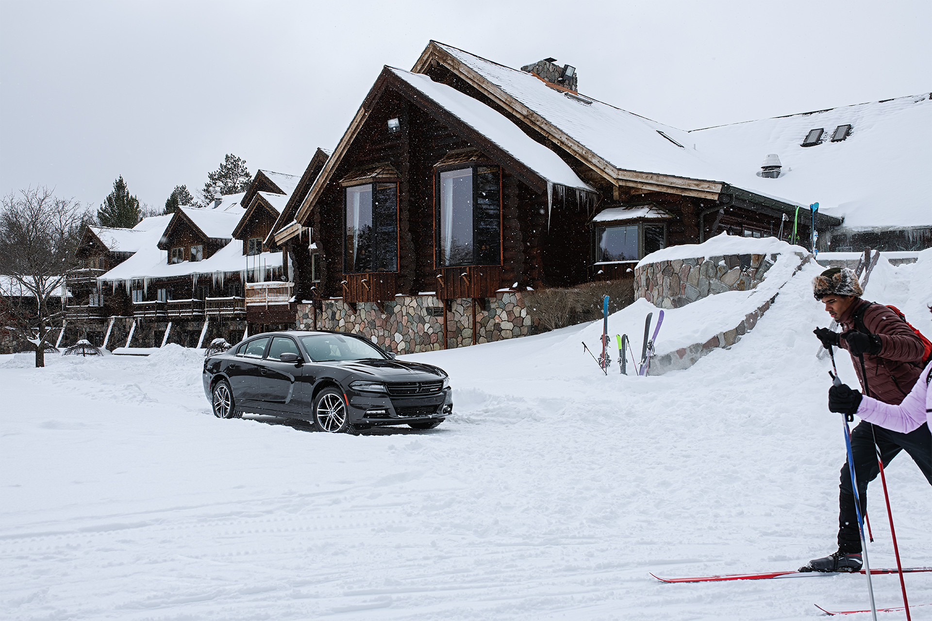La Dodge Charger 2020, illustrée en noir, stationnée dans l’entrée enneigée d’un chalet de ski, près d’un homme qui fait du ski