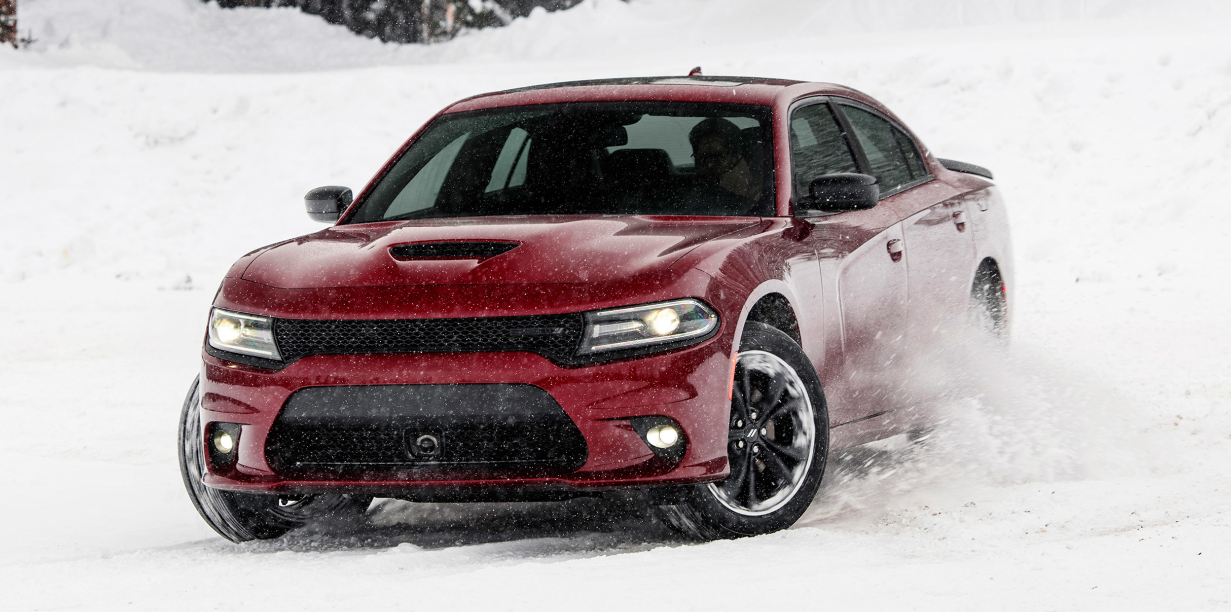 Dodge Charger bourgogne au bas d’une pente enneigée avec des arbres couverts de neige en arrière-plan.
