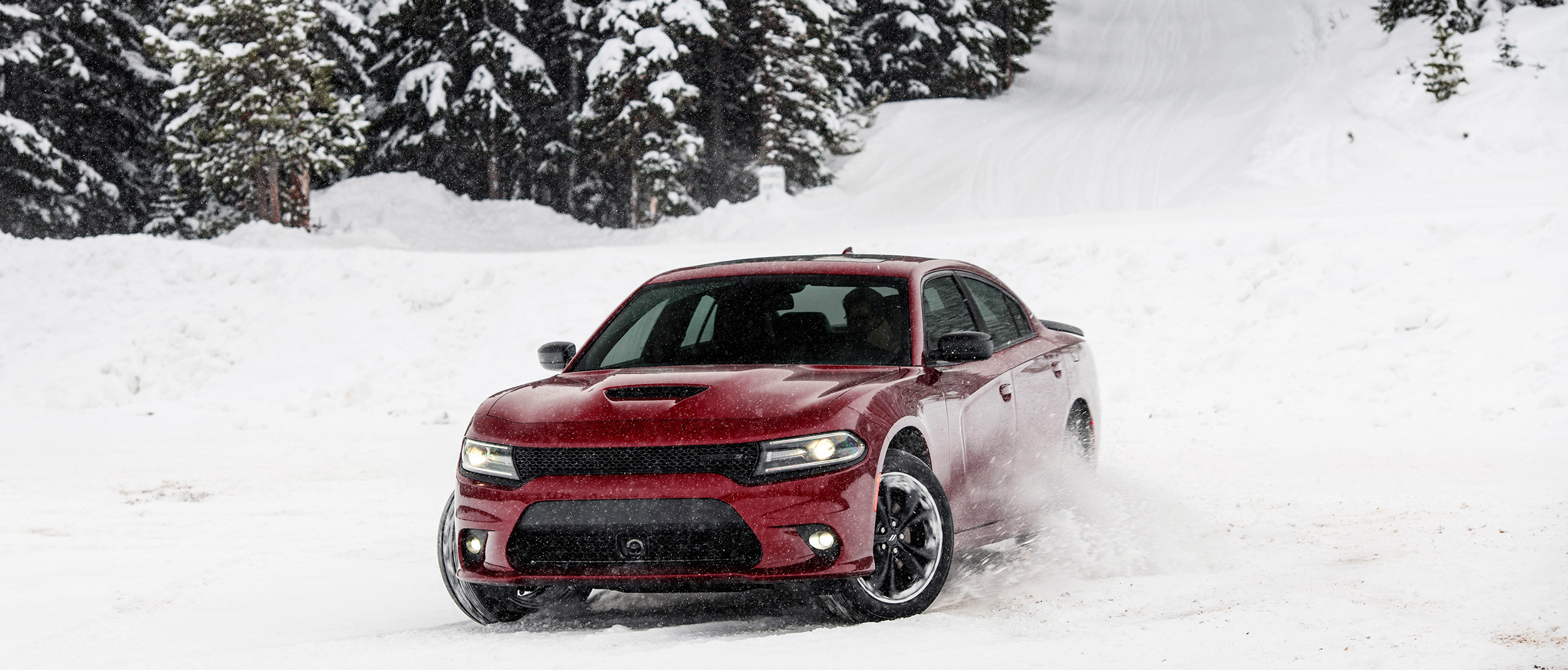 Burgundy Dodge Charger at the bottom of a snowy hill with snow-covered trees in the background.