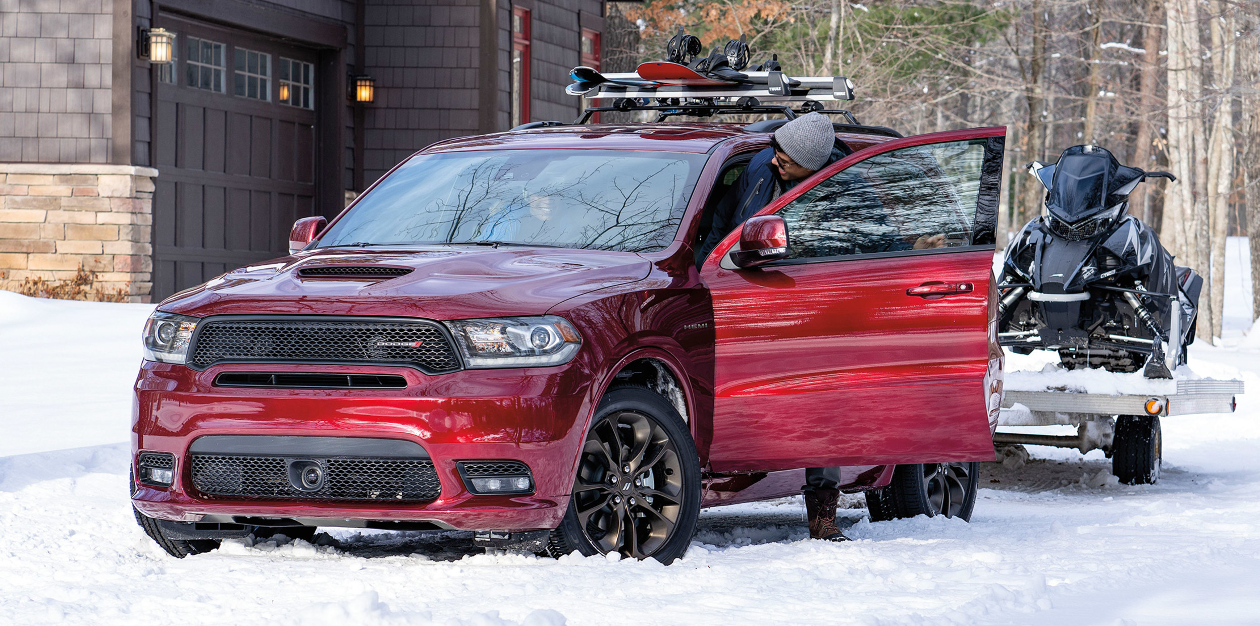 The driver steps out of a burgundy Dodge Durango parked by a home in the wilderness. There are skiis on the roof rack and a snowmobile hooked to the rear.