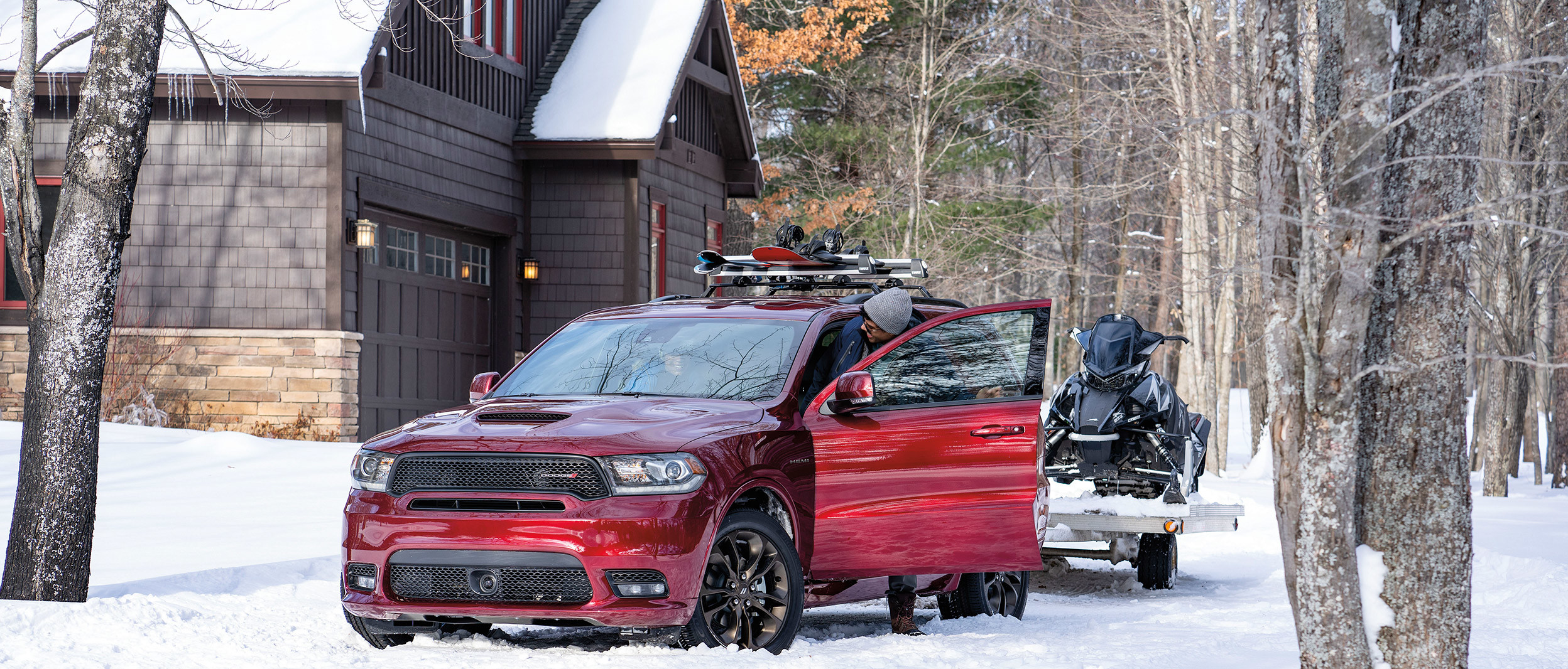 The driver steps out of a burgundy Dodge Durango parked by a home in the wilderness. There are skiis on the roof rack and a snowmobile hooked to the rear.