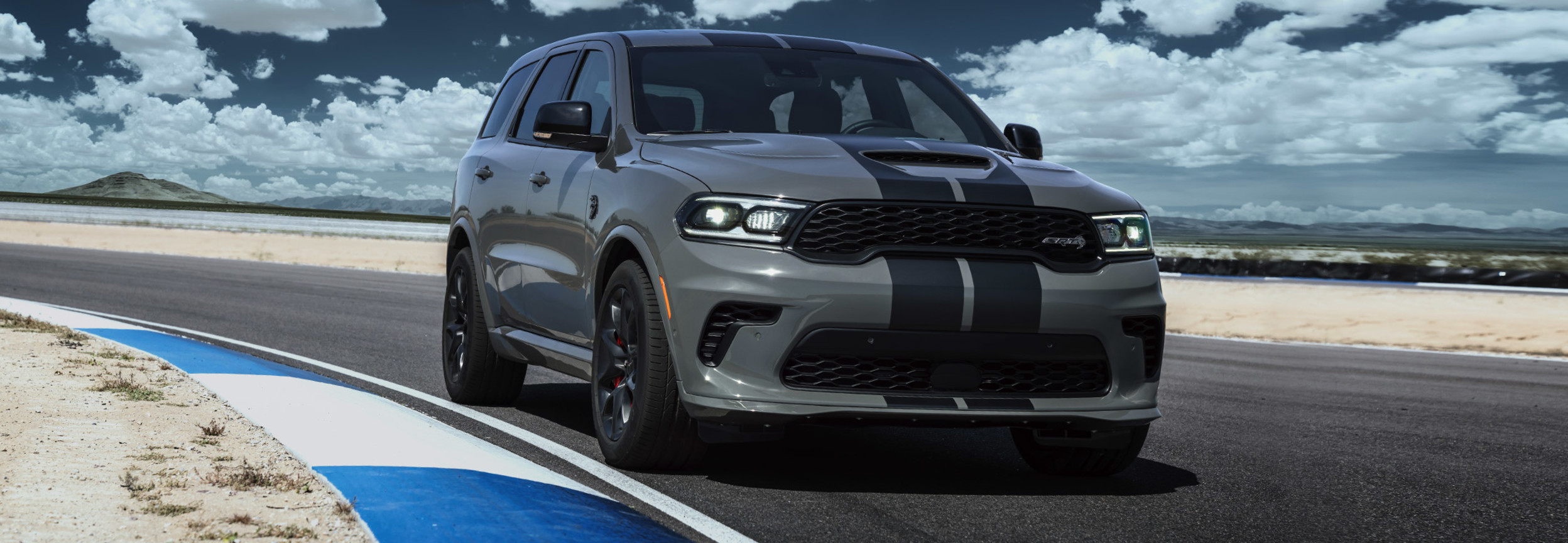 Front left-angle view of a grey Dodge Durango parked on the corner of a race track on a sunny day.