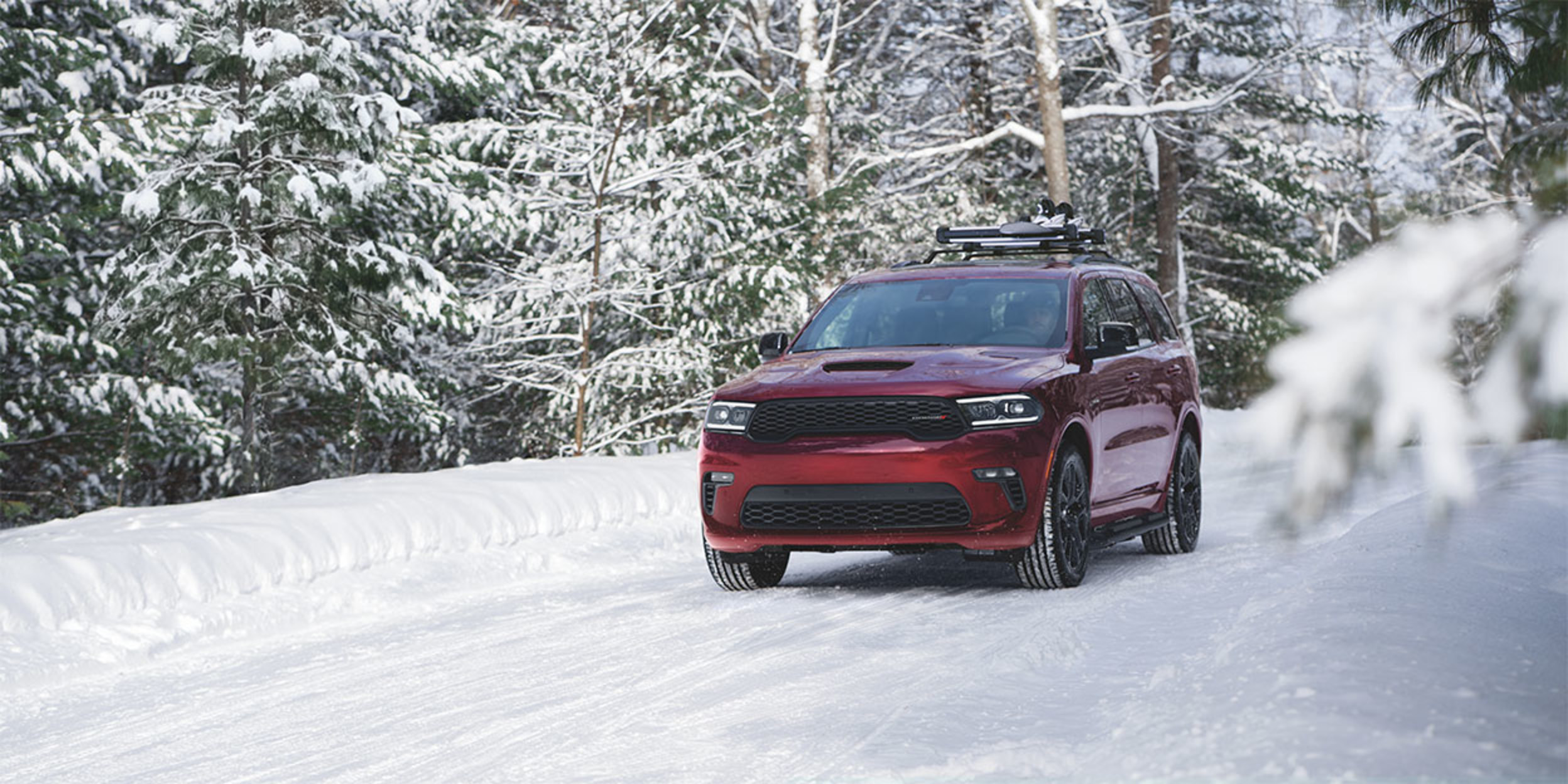 Front view of a red 2024 Dodge Durango shown driving on a snow covered path with pine trees shown behind.