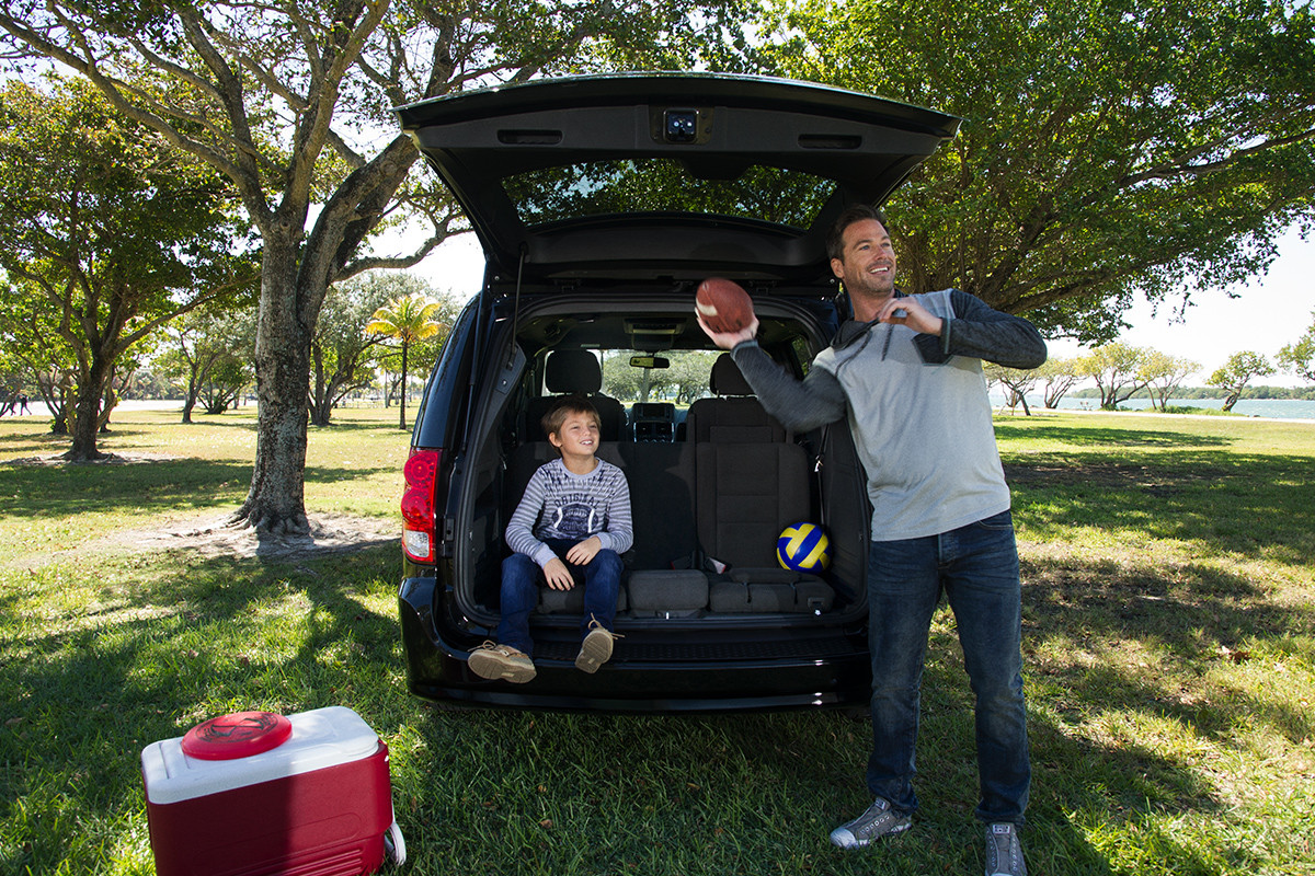 A boy sitting on the 3rd-row seat of the 2020 Dodge Grand Caravan with the tailgate open, while a man throws a football