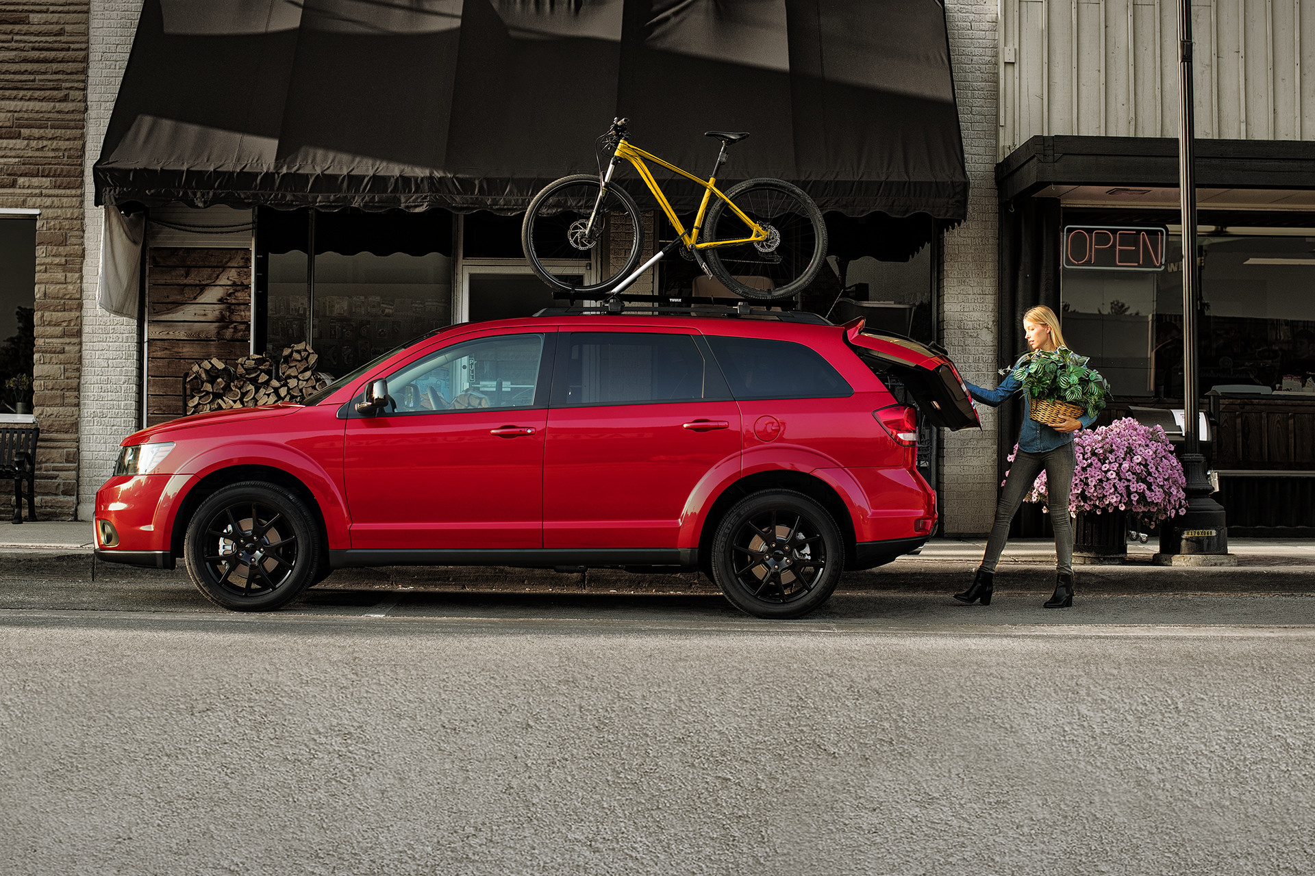 Red 2019 Dodge Journey with a yellow bike mounted on the roof, parked in front of a shop, with a woman holding a plant and opening the trunk.