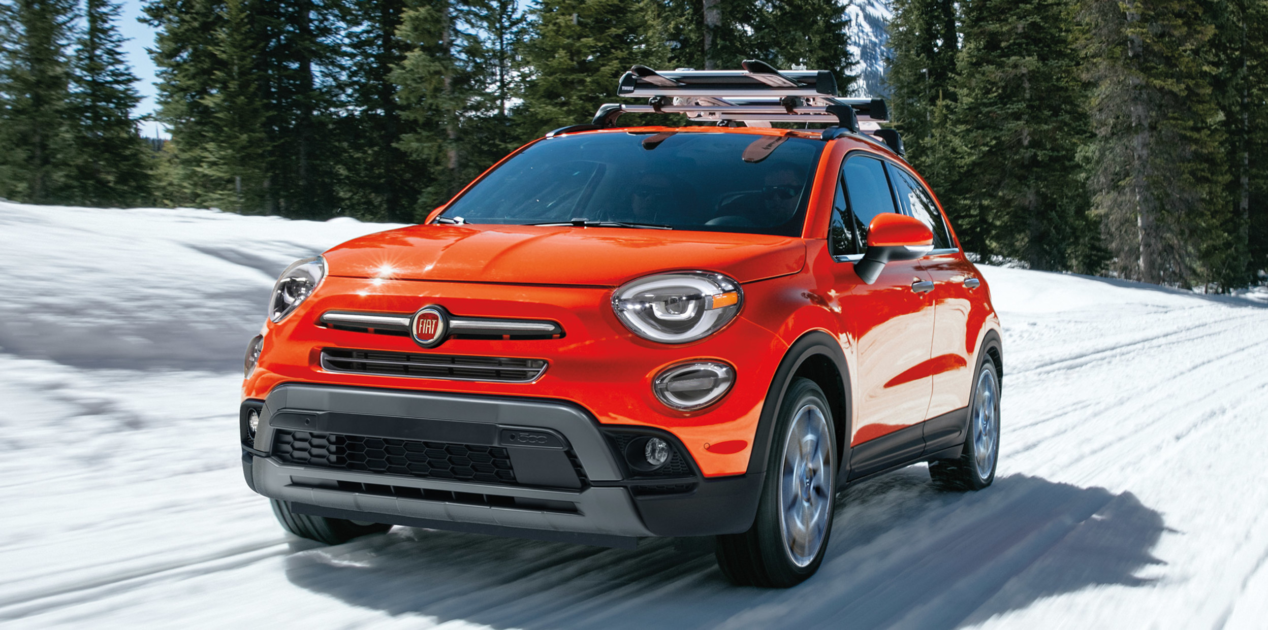 Red Fiat 500X driving on a snowy road with skiis affixed to the roof rack and mountains in the background.